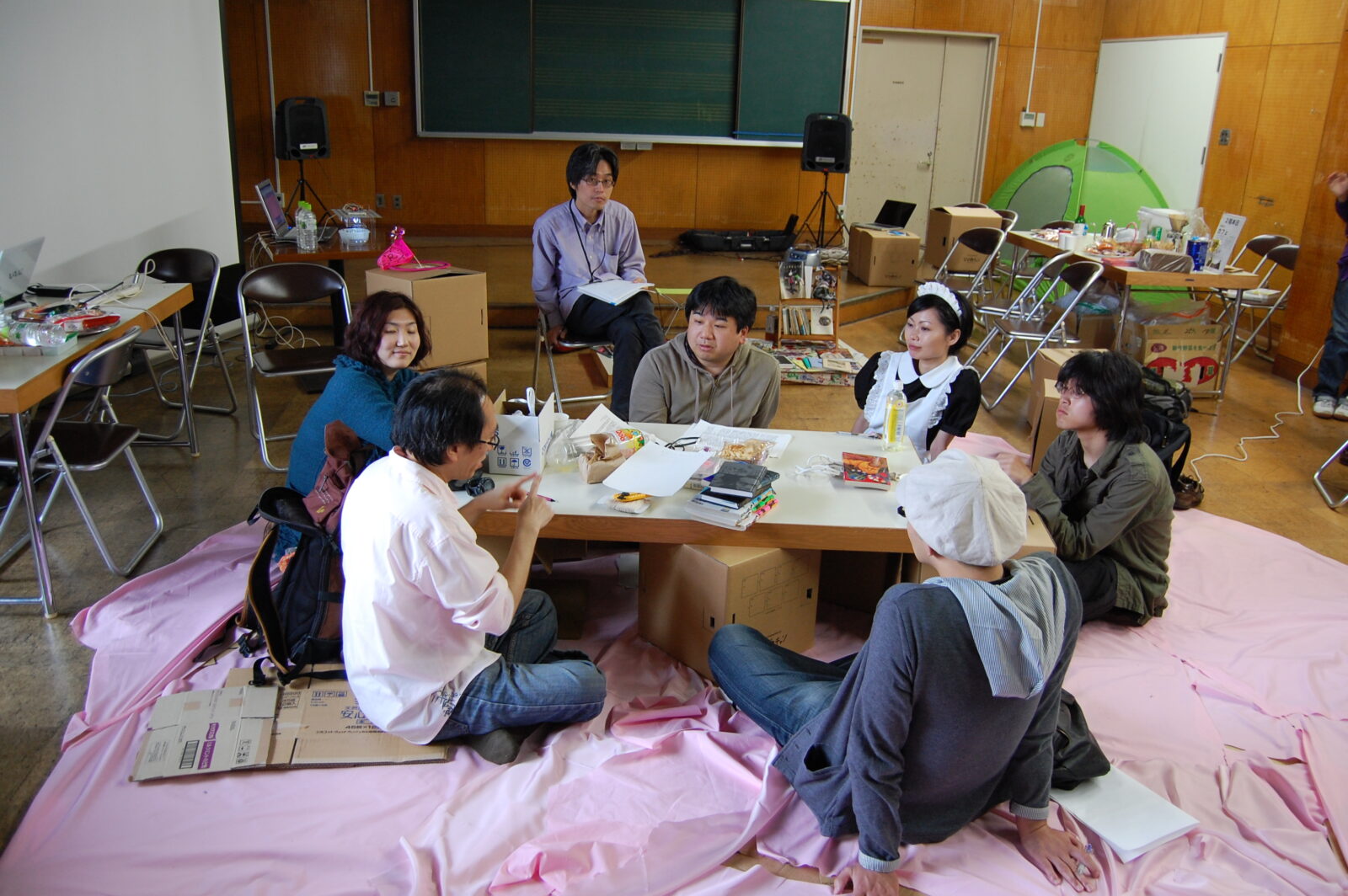 教室のような部屋の中、床の上に敷かれたピンクの布の上に座卓が置かれ、その周りに複数の参加者が座っている