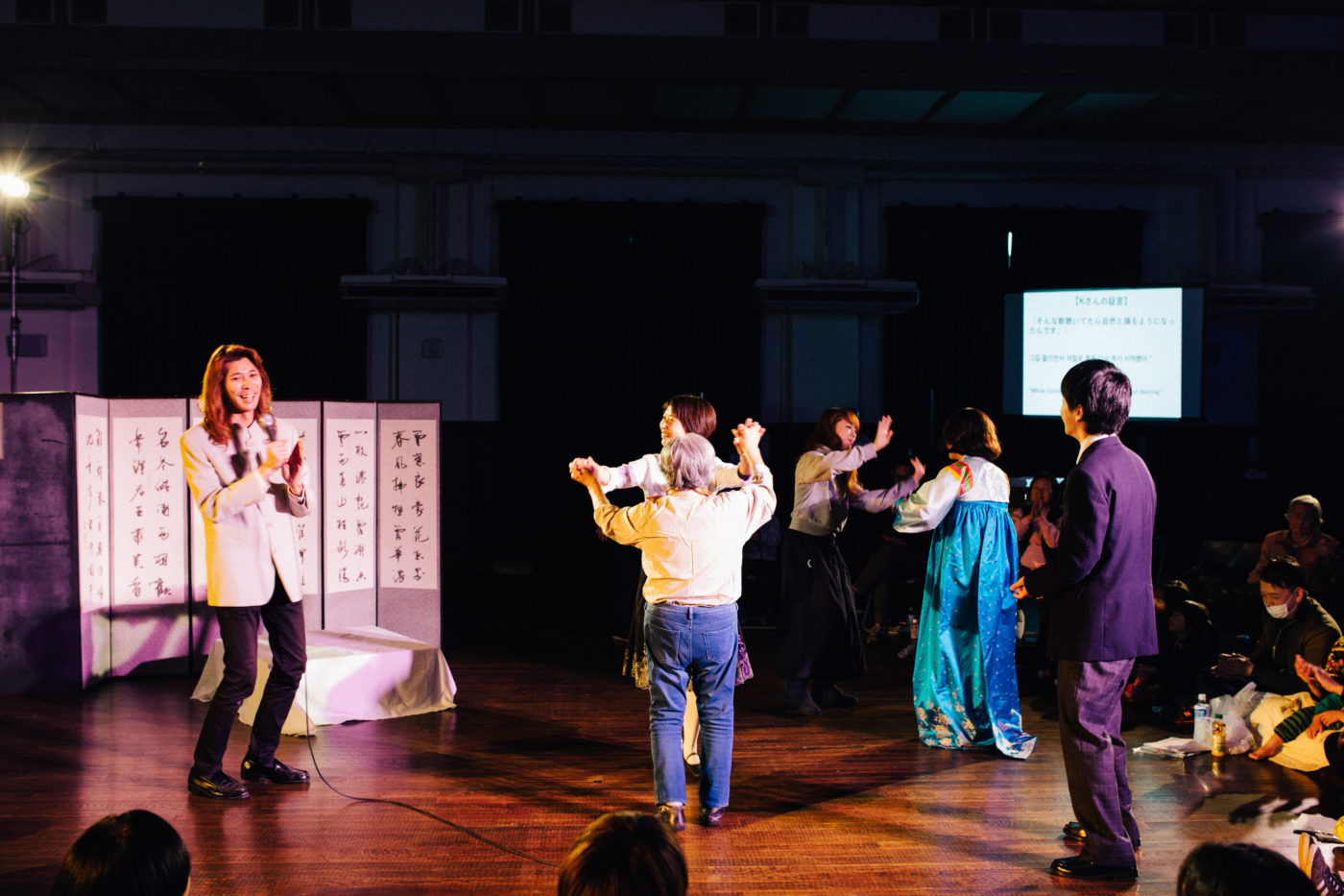 マイクを持った人がいて、その横で手を取り合ってダンスをしている人たちがいる風景。奥の人は民族衣装・チマチョゴリを着ている