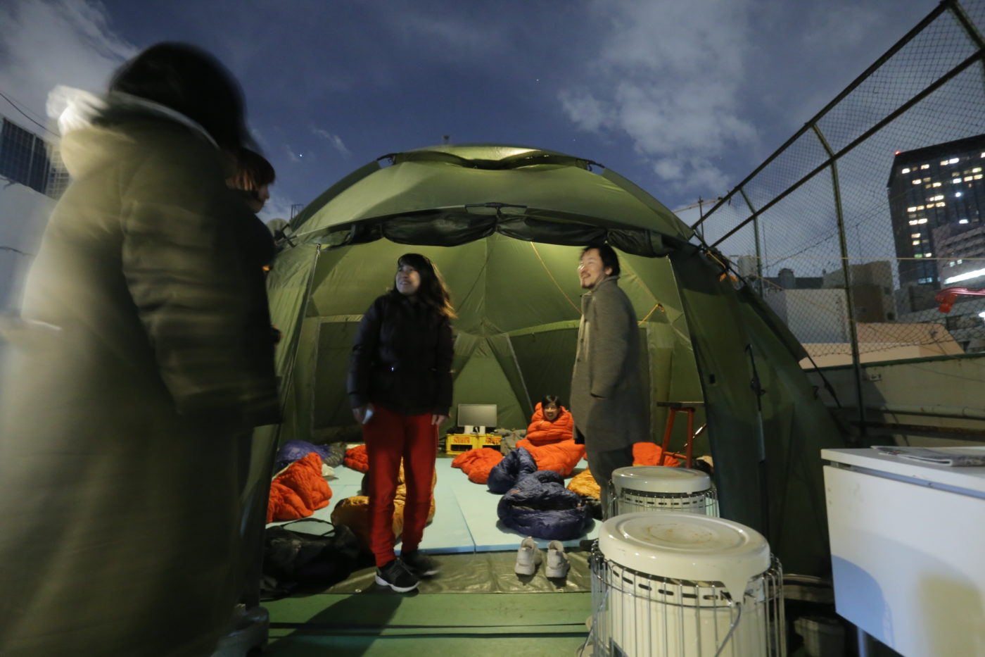 ビルの屋上にテントを貼って、そのなかにたくさんの寝袋が用意されている様子。その寝袋にくるまっている人がいたり、入り口で談笑したりしている人たちがいる