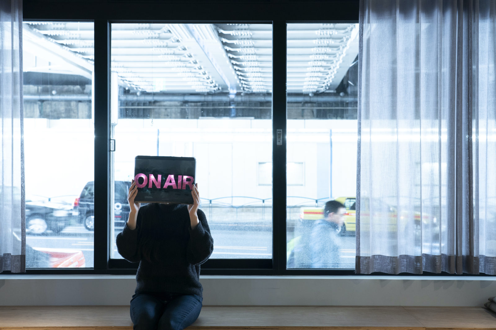 窓際に座った人がピンクの文字で「ON AIR」と表示されたデバイスを顔の前に掲げている風景。窓の向こうには道路があり、車が走ったり、人が歩いたりしている
