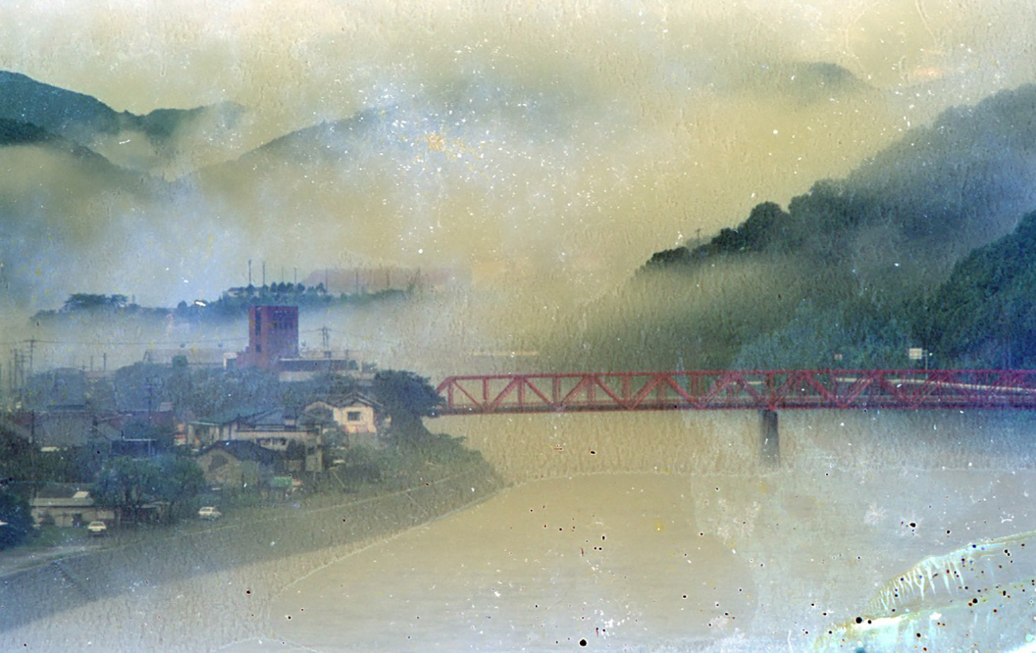 大きな川が流れていて、その奥には山々が連なっている。川の上には赤い橋が架かっていて、その先には住宅街がある風景