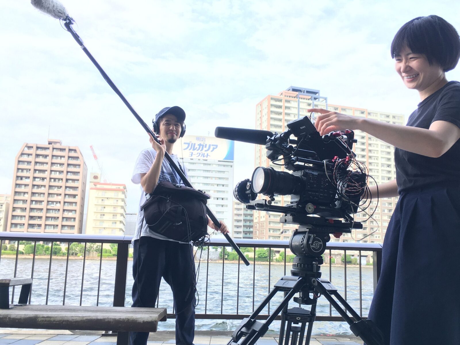 川を背に三脚を立て、カメラを構えている人と、音声マイクで音を拾っている人がいる。二人とも笑っている。川の向こう側には高いマンションが並んでいる