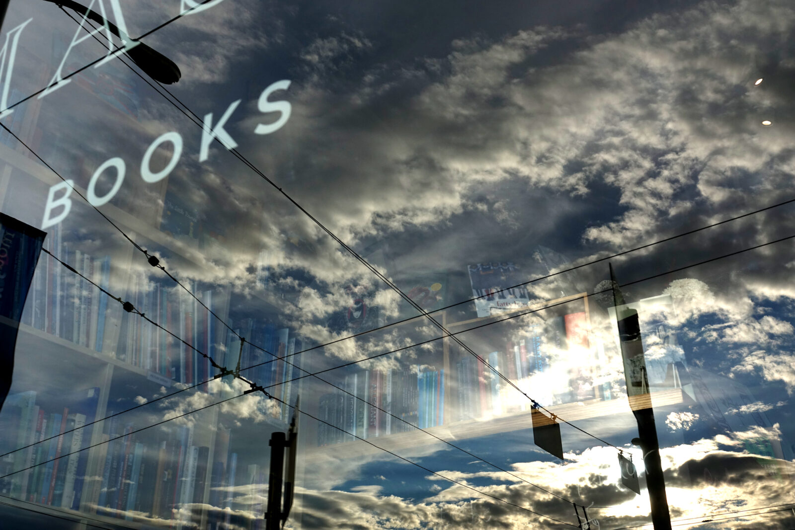 BOOKSという文字が書かれたガラス窓に電線や雲間から太陽が差し込んでいる風景が反射して写り込み、その奥には本棚が見えているイメージ写真