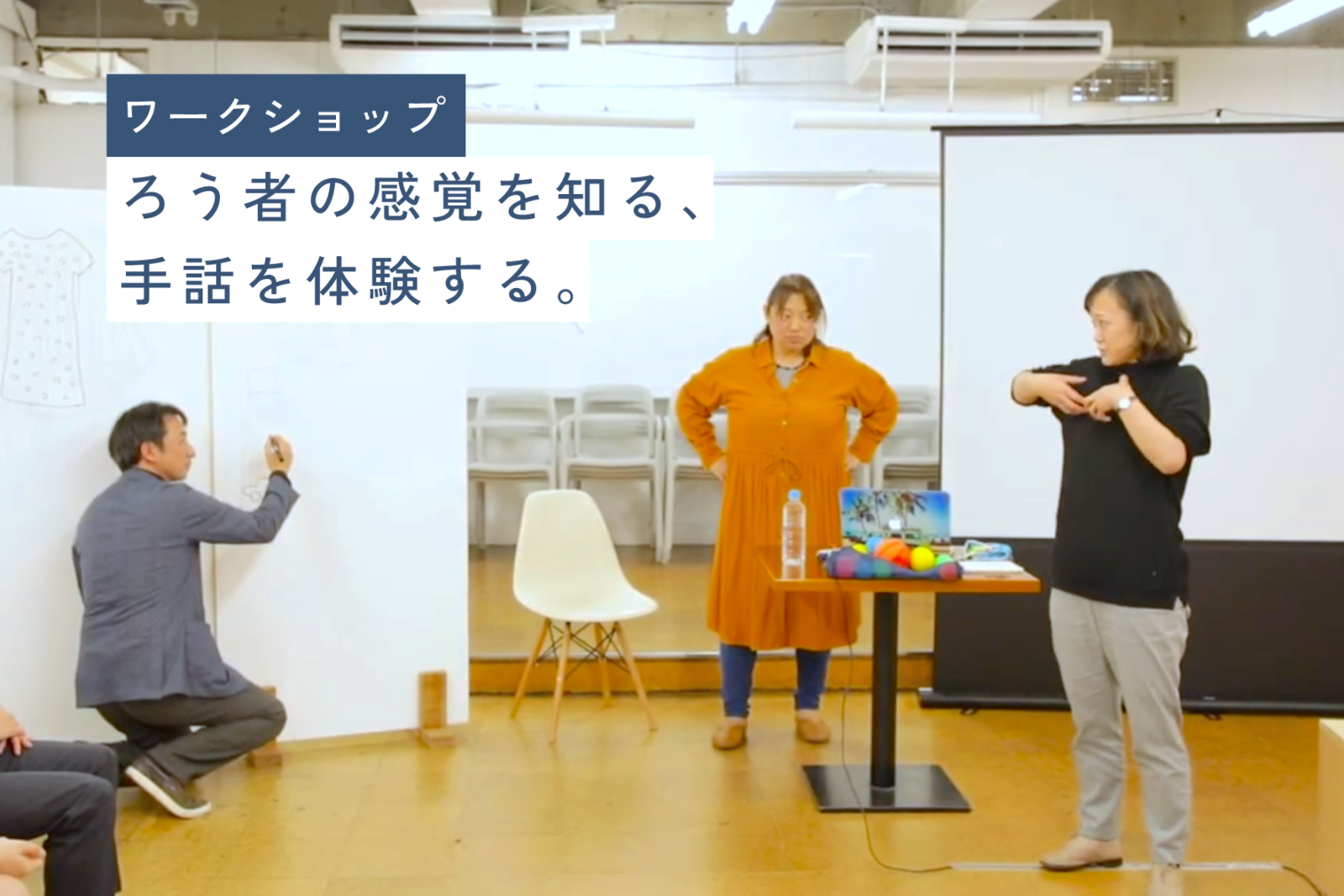 「ワークショップ　ろう者の感覚を知る、手話を体験する。」という文字の背景に、手話をしている人がいて、そこから受け取ったイメージをホワイトボードに絵で描いている人がいるイメージ画像