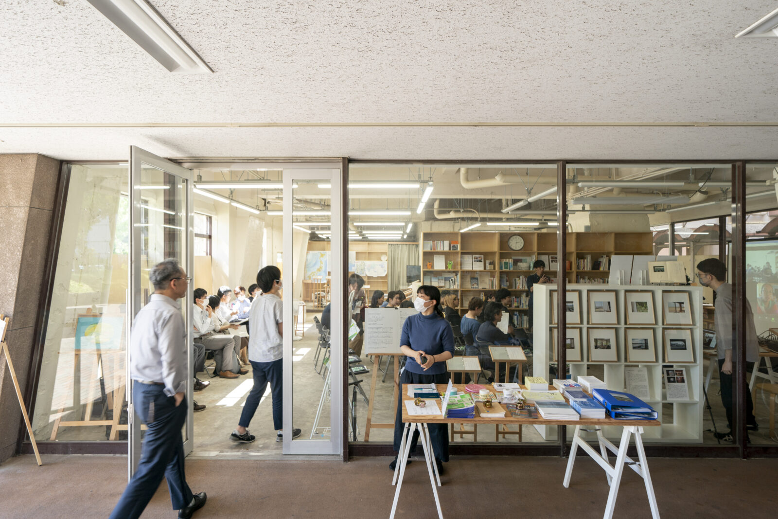 ガラス張りの一室の前に本の並んだ机が置かれている。ガラス張りの部屋の中には本棚があり、部屋の外と中には数人が歩いたり立ったりしている。