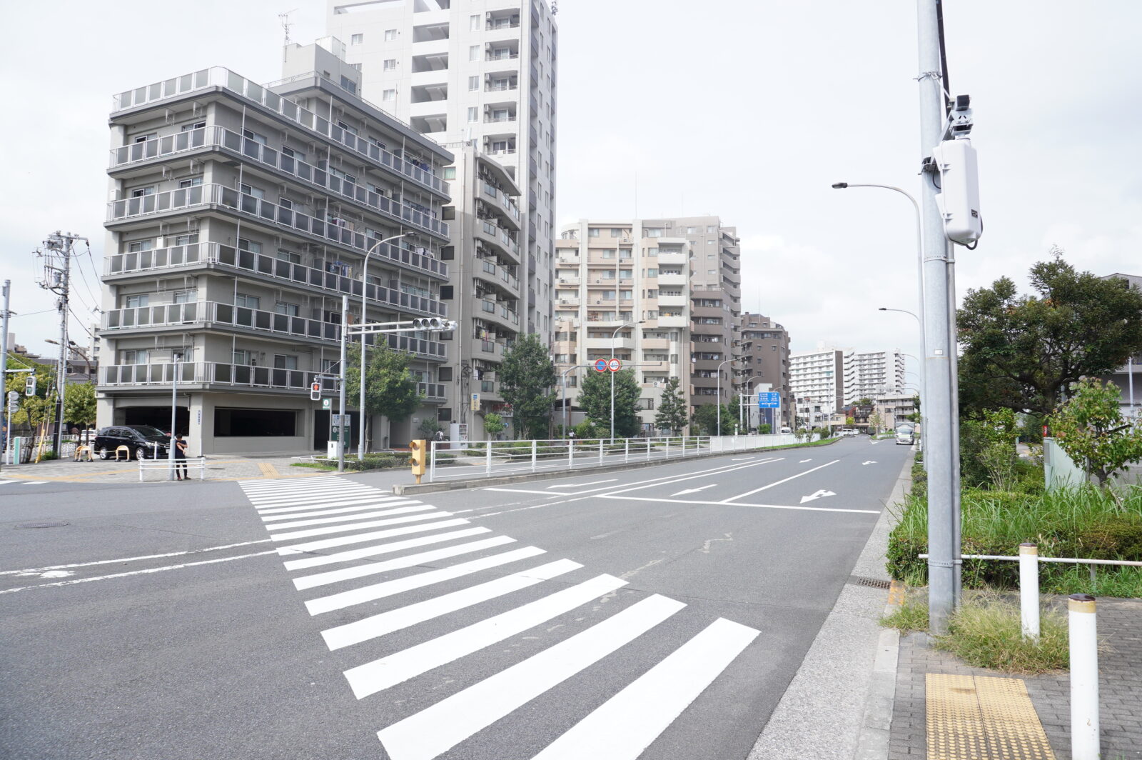 大きな幹線道路に横断歩道がわたっている。道路の向こうには、6階建てのマンションや高いビル、団地などが並んでいるのが見える