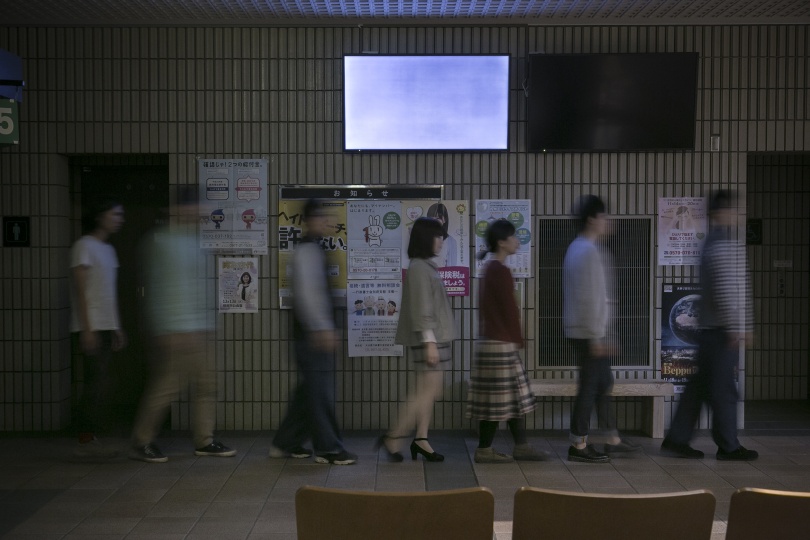 薄暗い通路を、7人ほどの人が一列になって歩いている。壁には様々なポスターが掲示されている