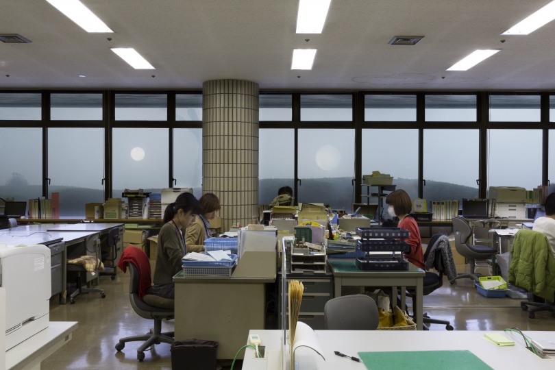 たくさんの机やコピー機、資料などが置かれた空間のなか、4人ほどの人物が座って仕事をしている。一面が窓になっており、窓の外には山や光る球体が見える