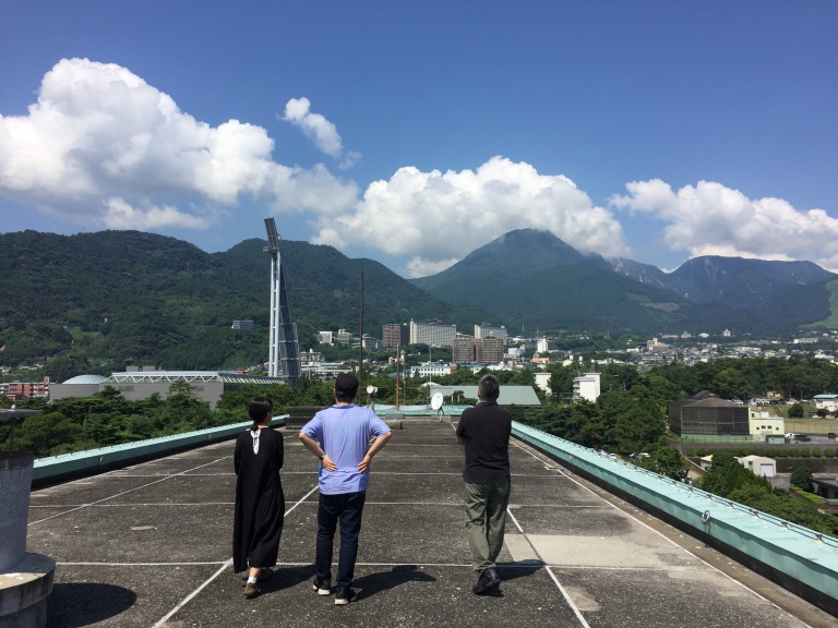 建物の屋上を3人の人物が歩いている。屋上からは空や山、街並みが見える