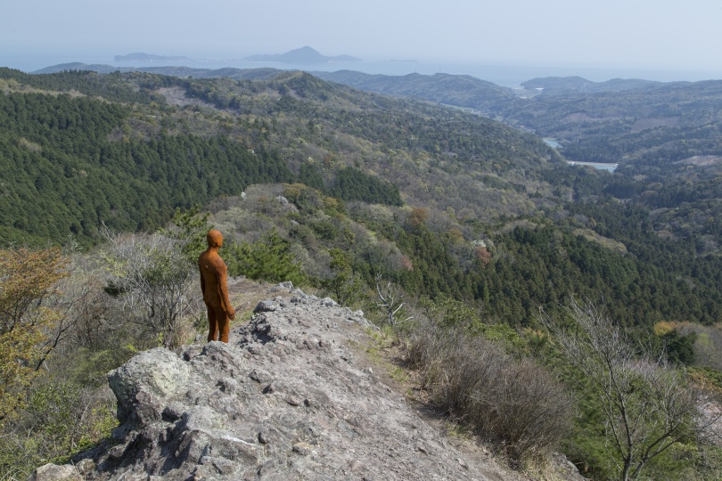 岩肌の見える山頂に、黄土色で人型の彫刻作品が置いてある。周囲は山々に囲まれている