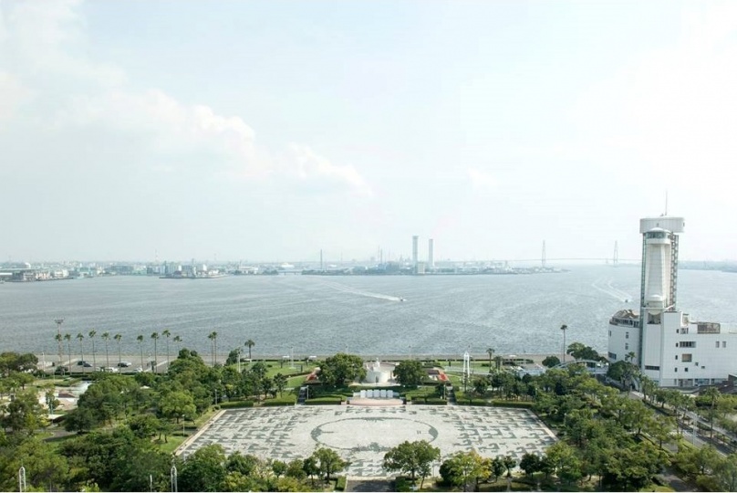 陸地に沿って、手前には緑豊かな公園があり、奥には建物が密集している港がある。港の中には船が一隻走っており、水しぶきを上げている