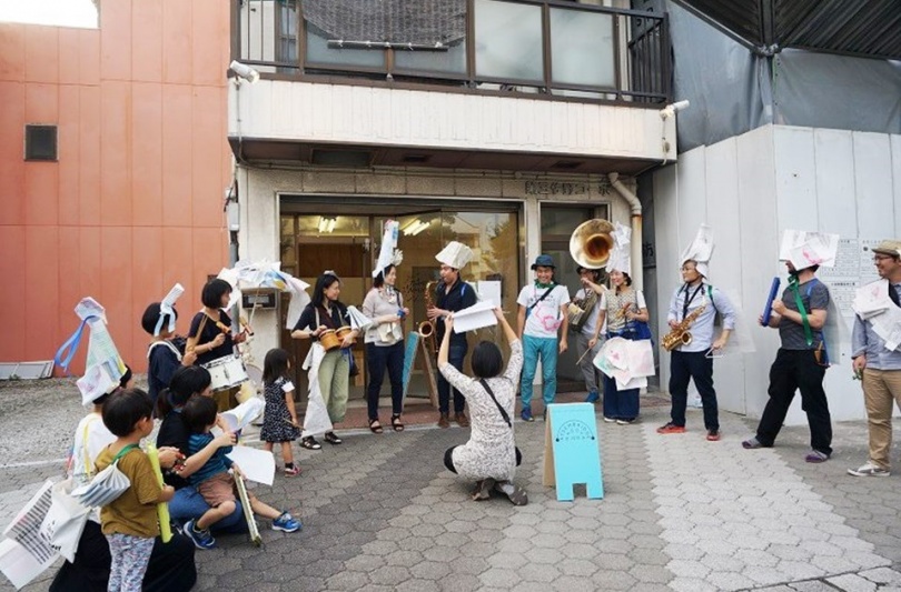 建物の前の路面に、20人ほどの人物が半円状になって並んでいる。それぞれ被り物をしたり楽器を持ったりしている。人々の真ん中にはしゃがんでいる人物がおり、頭の上に紙を掲げて何か合図をしている