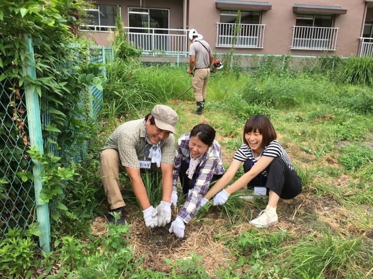 野草が生い茂った広場で草取りをする人々。手前の3人は笑いながら、手作業で草を抜いている。奥側には草刈り機を使って作業する人物が1人いる