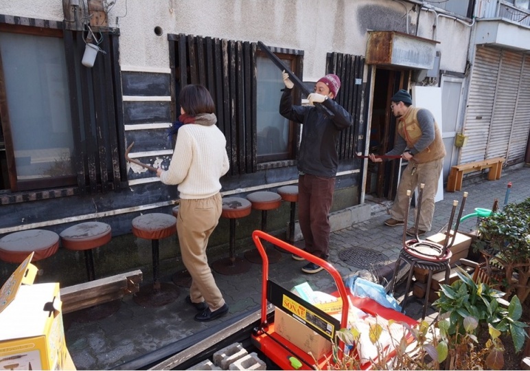 路地に3人の人物が並び、建物の窓の枠を壊すなどの作業を行っている。3人の周囲にはほこりをかぶった椅子や、段ボール箱、台車などが置いてある