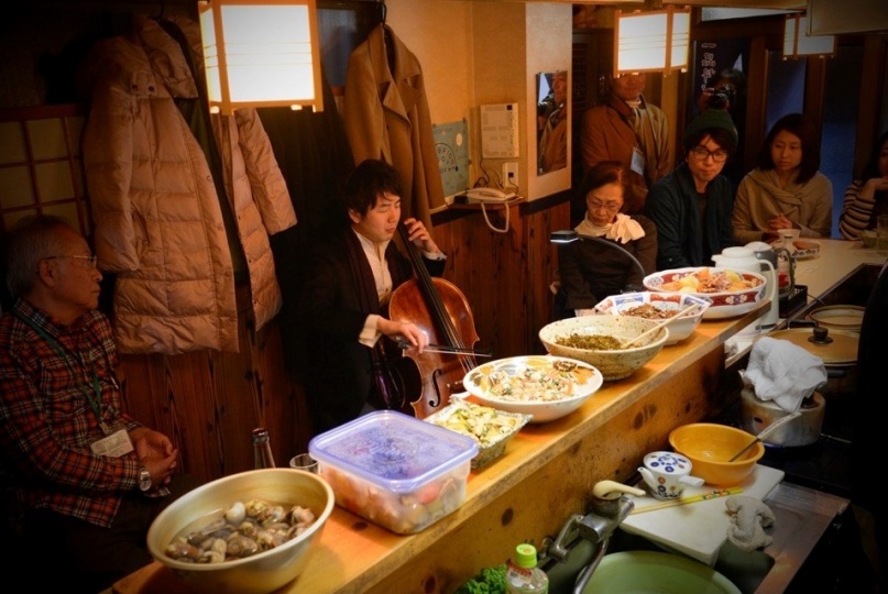 カウンター式の飲食店で、客席側の真ん中あたりにチェロ奏者が座って演奏している。その周りに複数の観客が座って演奏を聴いている