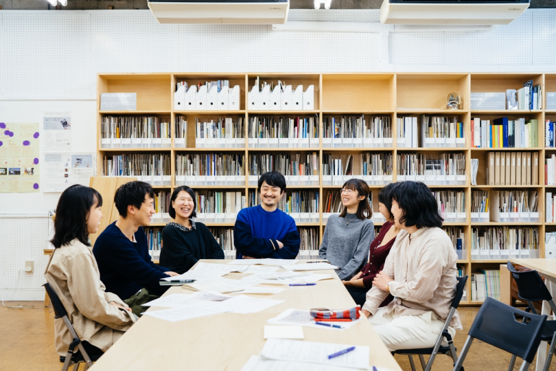 学校の教室のような部屋の中、壁に大きな本棚が設置されている。その前に7人のメンバーが机を囲んで座り、笑いながら話している